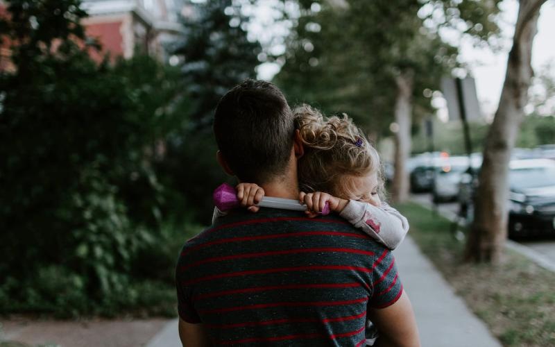 Close to the center of everything tired little girl rests her head on her father's shoulder as he carries her down the city sidewalk